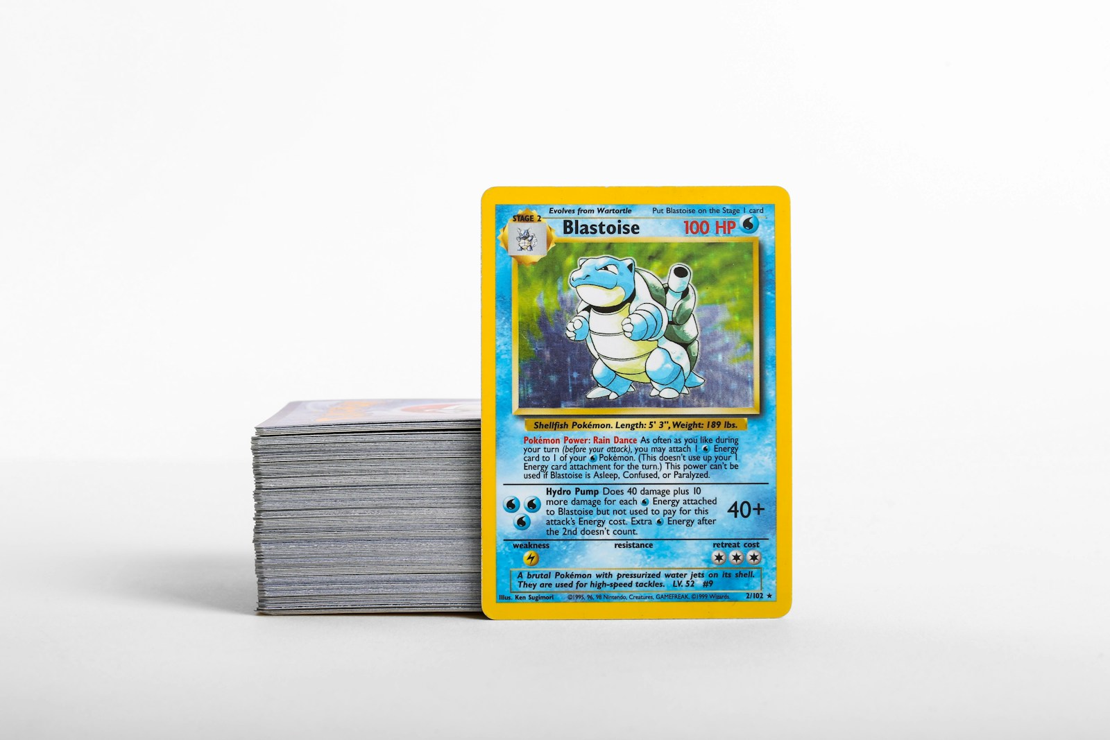 A Blastoise Pokémon card standing beside a stack of trading cards on a light surface.